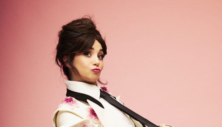 Een vrouw met donker opgestoken haar en opvallende make-up poseert tegen een roze achtergrond.