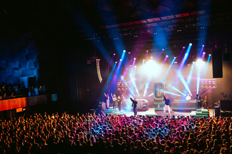 Een groot publiek bij een concert met felgekleurde podiumlichten.