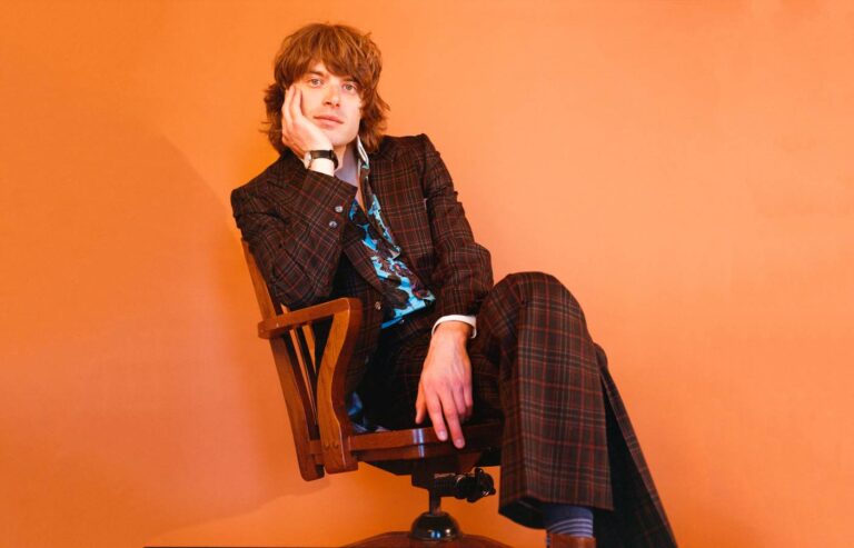 Een persoon in een donker pak zit op een houten stoel voor een fel oranje achtergrond.