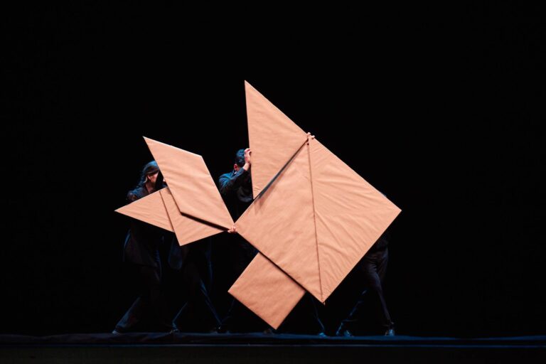 Meerdere grote oranje origami-achtige panelen staan in een sculpturale compositie tegen een zwarte achtergrond.