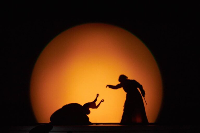 Twee silhouetten van mensen op een podium voor een grote oranje zon op de achtergrond.