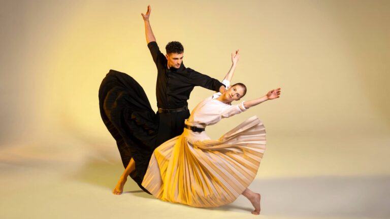 Twee balletdansers in een dynamische pose, één in een zwarte outfit en de andere in een gele jurk, tegen een lichte achtergrond.