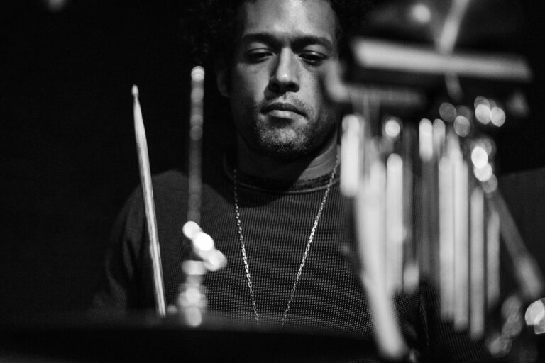 Zwart-wit foto van een mannelijke drummer met twee drumsticks in de voorgrond.