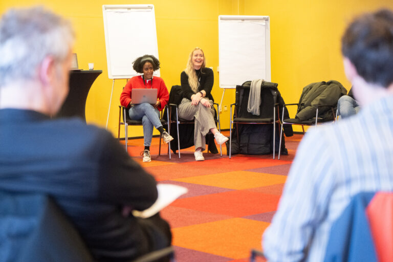 Drie sprekers zitten op stoelen voor een publiek in een felgele zaal met flipovers op de achtergrond.
