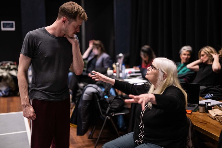 Een blonde vrouw wijst naar iemand terwijl een man naast haar naar beneden kijkt, met meerdere mensen zittend op de achtergrond in een theaterachtige ruimte.