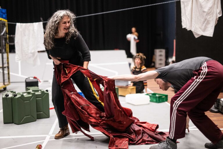 Een vrouw met lange krullende grijze haren knielt op een theaterpodium in een lange rode jurk.