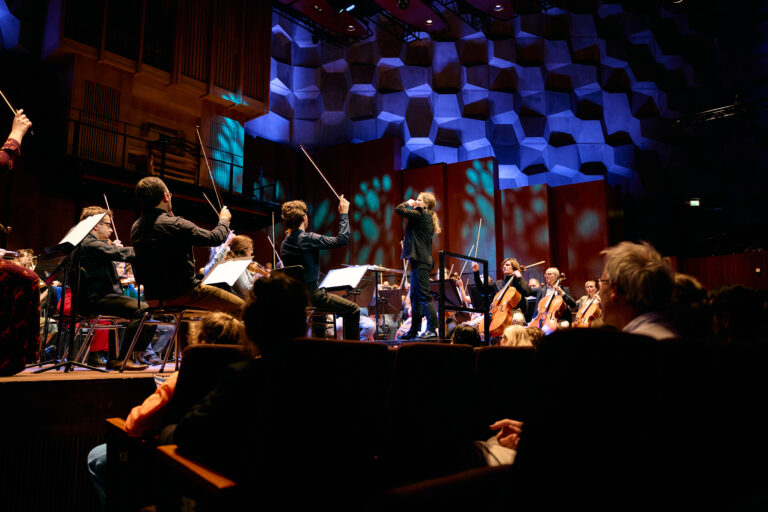 Een symfonieorkest speelt op een podium onder sfeervolle zaalverlichting, geleid door een dirigent.