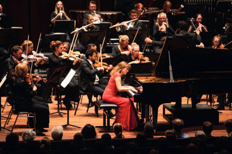 Een pianiste in een rode jurk speelt samen met een groot orkest op een concertpodium.