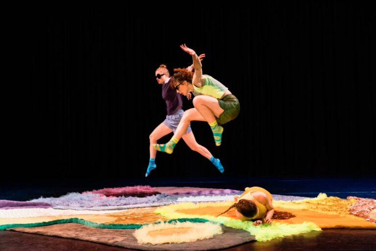 Twee dansers springen energiek boven een kleurrijke, zachte ondergrond terwijl een derde danser op de grond ligt.