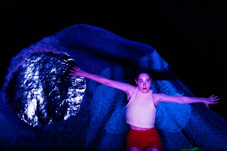 Een persoon ligt horizontaal onder een grote blauwe doek tegen een donkere achtergrond.