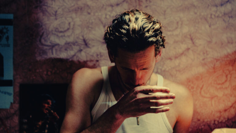 Man met kort donker haar, in een wit tanktop, buigt naar voren en houdt zijn hand voor zijn mond.