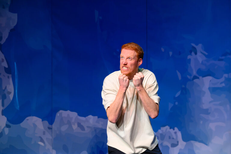 Een man in een wit shirt staat op een podium tegen een achtergrond van een blauwe lucht met wolken.