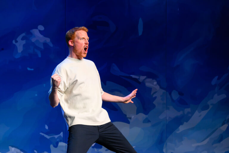 Een man in een wit T‑shirt en donkere broek danst op een podium tegen een blauwe achtergrond.