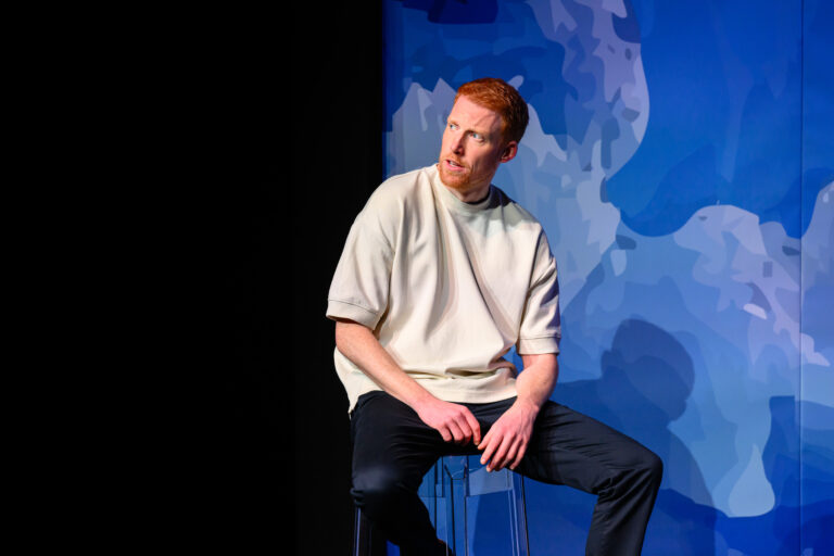 Een man zit op een kruk voor een blauwe achtergrond met een wereldkaart-achtig patroon.