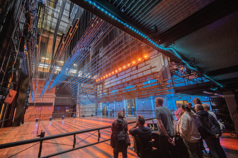 Industriële binnenruimte met staalconstructies en blauwe en oranje verlichting, terwijl een paar mensen langs een leuning staan.