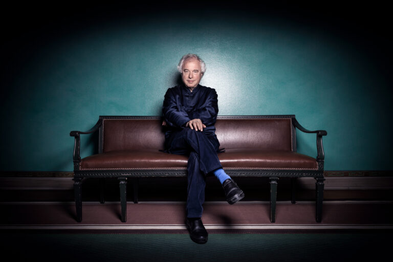 Een persoon in een donker pak zit op een leren bank tegen een turquoise achtergrond.