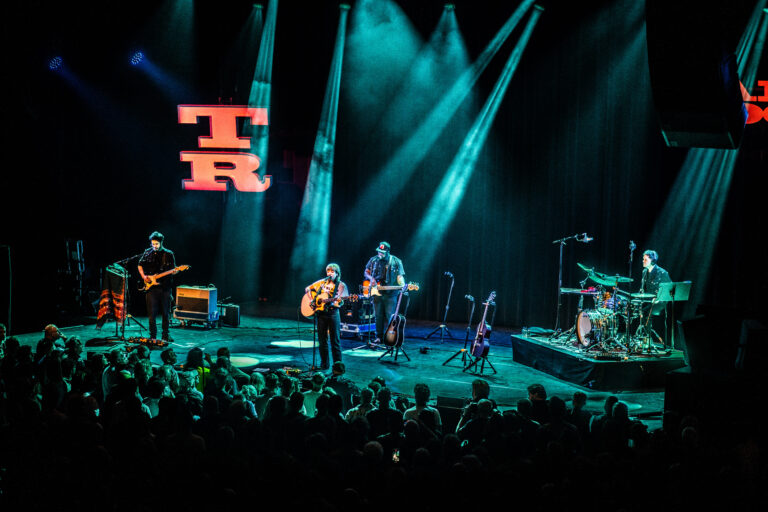 Een band speelt live op een podium met blauwe lichtstralen terwijl het publiek toekijkt.