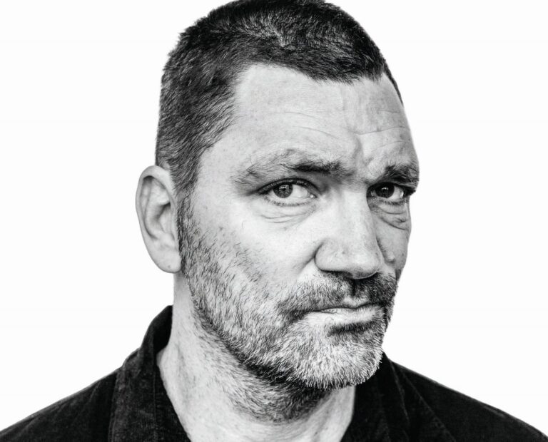 Zwart-wit portret van een man met korte haren en een stoppelbaard, die recht in de camera kijkt.