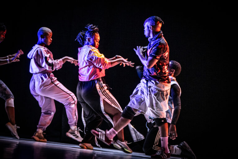 Een groep dansers in witte en rode outfits voert een dynamische choreografie uit op een donker podium.