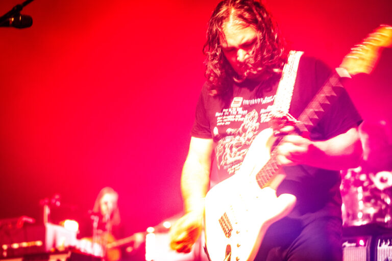 Een gitarist met lang haar speelt een witte elektrische gitaar onder fel rood podiumlicht.