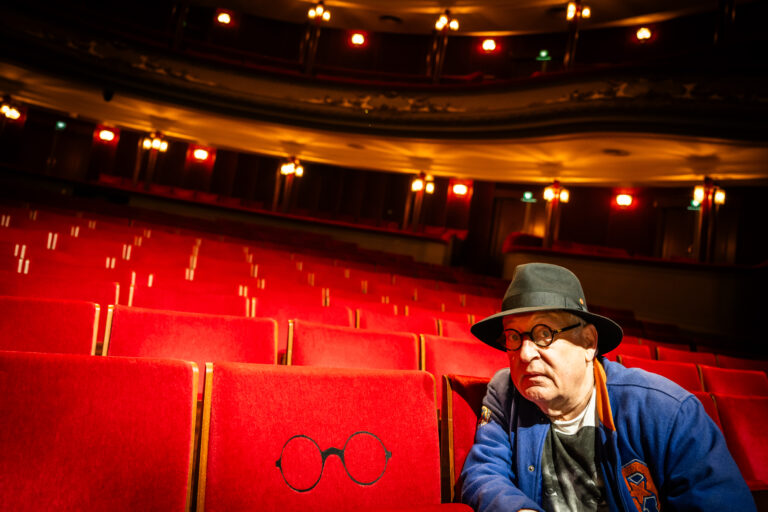 Een man met hoed en bril zit in een zaal met rode theaterstoelen.