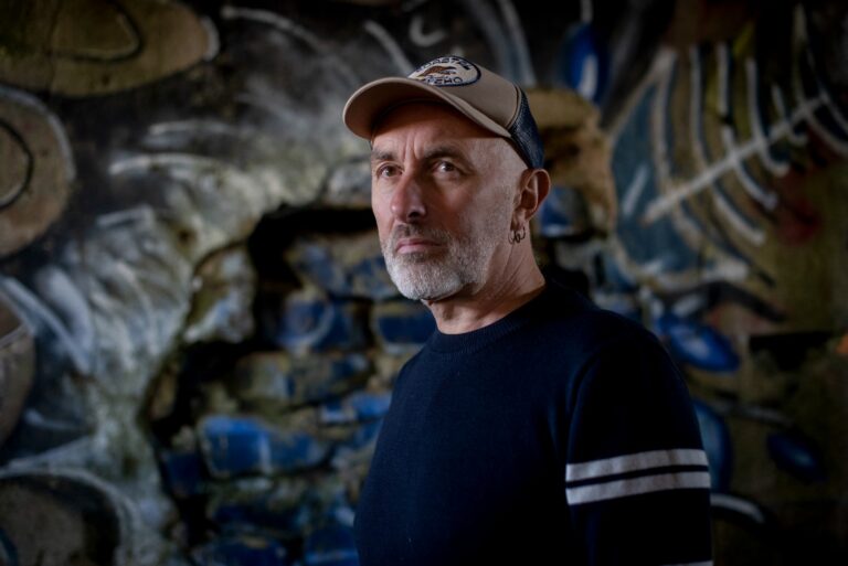 Een man met een pet en donkere trui staat voor een muur vol graffiti.
