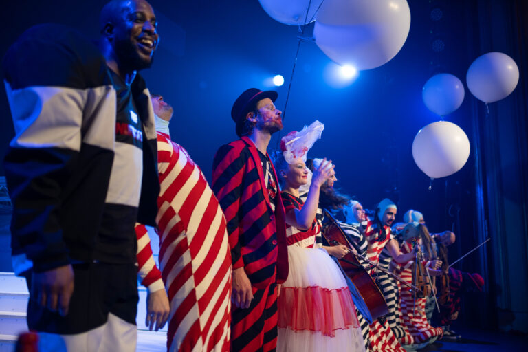 Een groep performers op een podium in kleurrijke kostuums, met ballonnen op de achtergrond en blauwe verlichting.