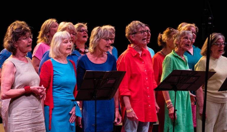 Een vrouwenkoor zingt op een podium, gekleed in felgekleurde jurken.