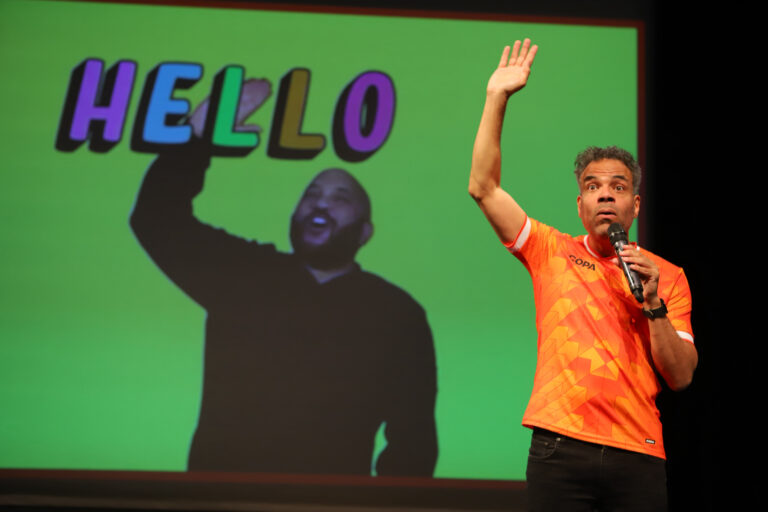 Twee mensen staan op een podium; één is een donker silhouet en de ander draagt een oranje shirt met een arm omhoog tegen een groene achtergrond.