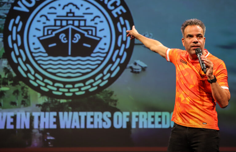 Een sporter in een oranje shirt staat voor een groot digitaal scherm met een ronde blauwe embleem en de tekst “The Waters of Freedom”.