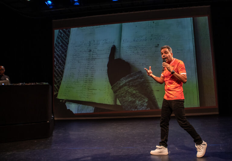 Een spreker in een fel oranje shirt staat op een podium en geeft een presentatie, met een grote schermachtergrond achter hem.