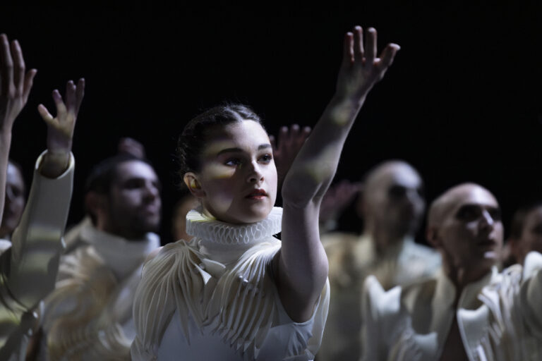 Een danseres in een wit kostuum steekt haar hand omhoog, omringd door andere dansers in een donkere setting.