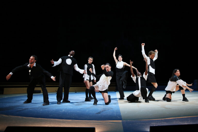 Een groep dansers in zwart-witte kostuums voert een dynamische choreografie uit op een podium.