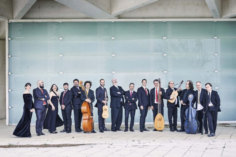 Een groep muzikanten in formele kleding poseert buiten voor een glazen muur, met strijkinstrumenten zoals cello en violen.