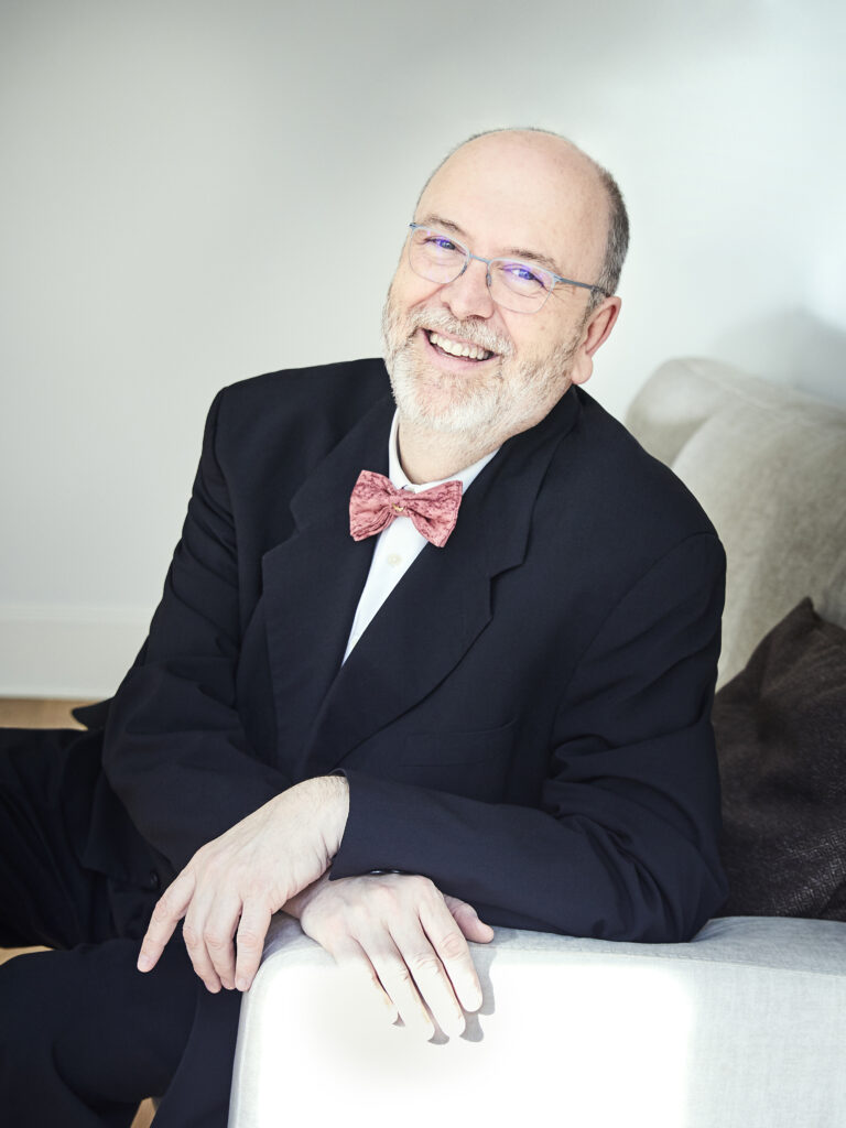 Een glimlachende man in een zwart pak en een vlinderdas, zittend op een bank.