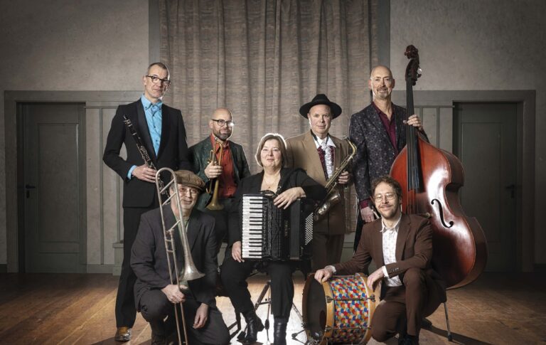 Een groep muzikanten poseert samen met verschillende instrumenten, waaronder een accordeon, viool, gitaar en bas.