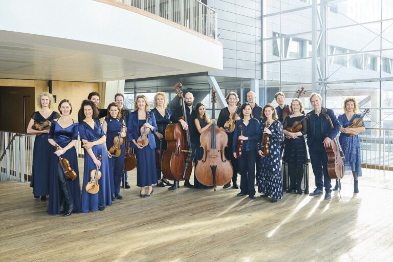 Een klassiek orkest poseert in een moderne zaal, gekleed in blauwe outfits met verschillende instrumenten in de hand.