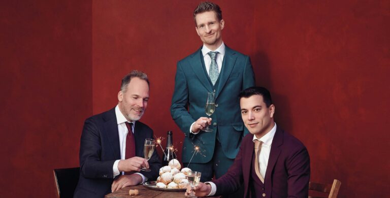 Drie mannen in pak staan en zitten aan een tafel met een taart in het midden tegen een rode achtergrond.