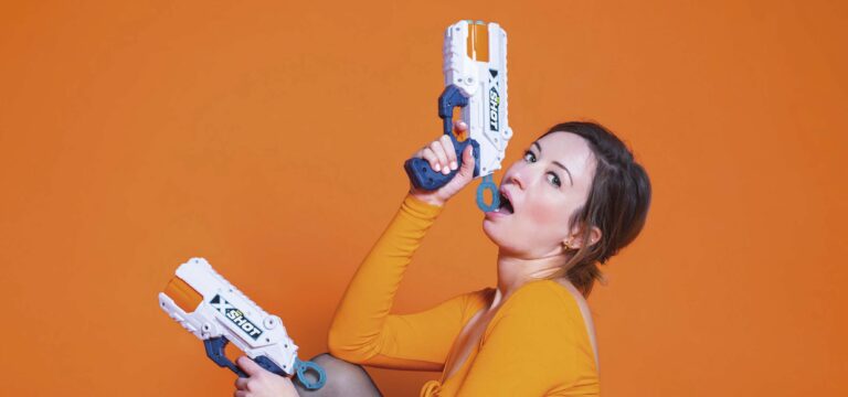 Een persoon in een oranje outfit richt een futuristisch blauw-wit speelgoedpistool omhoog tegen een oranje achtergrond.
