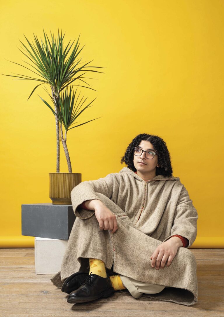 Een persoon in beige kleding zit op de vloer voor een felgele muur, met een hoge plant op de achtergrond.