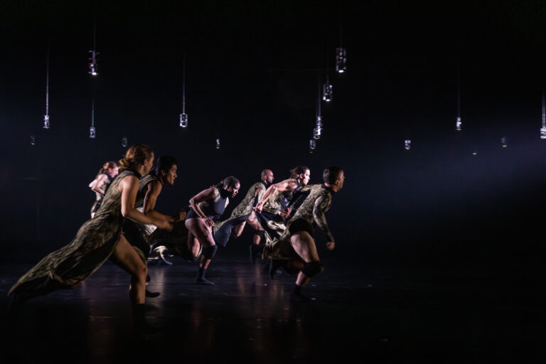 Een groep dansers in beweging tijdens een optredenscène, omringd door hangende lichten in een donkere ruimte.