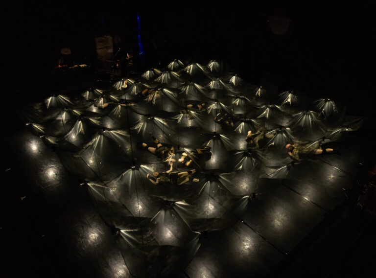 Een bovenaanzicht van een donkere ruimte met een netwerk van oplichtende tenten of zeilen, waar mensen onder liggen.