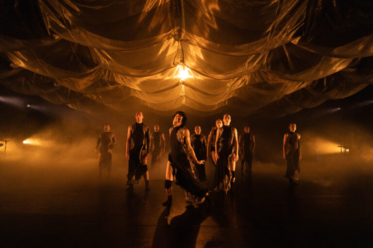 Een groep dansers presenteert een choreografie onder een sfeervolle, met licht en mist verlichte ruimte.