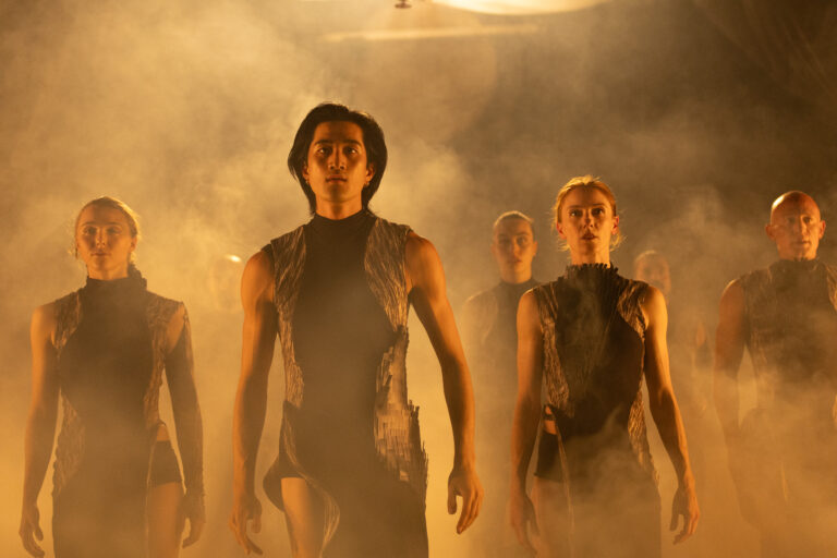 Een groep dansers in een rokerige omgeving, nauwe, donkere outfits dragend, loopt geconcentreerd naar voren.