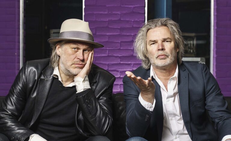 Twee mannen met lang haar zitten naast elkaar voor een paarse muur; de linkerman draagt een hoed en steunt zijn hoofd op zijn hand terwijl de rechter een open handgebaar maakt.