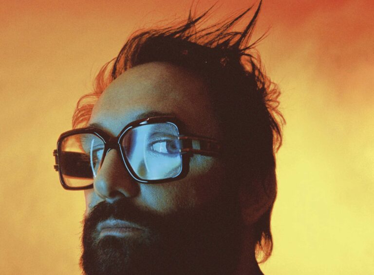 Een man met een baard en een vierkante bril tegen een oranje achtergrond.