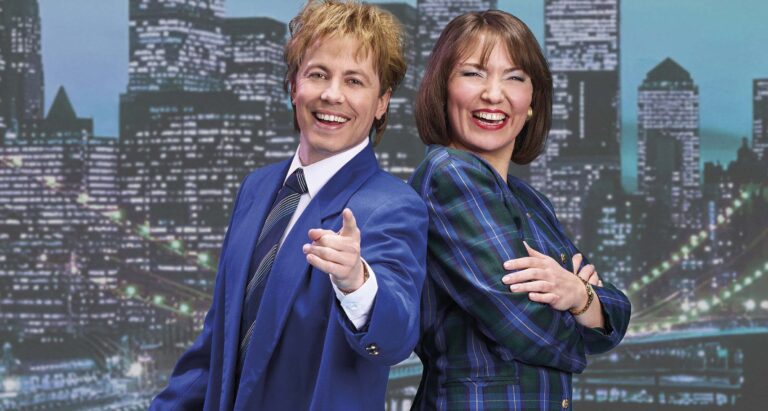 Twee mensen in zakelijke kleding glimlachen naar de camera, terwijl de man in een blauw pak naar de kijker wijst.