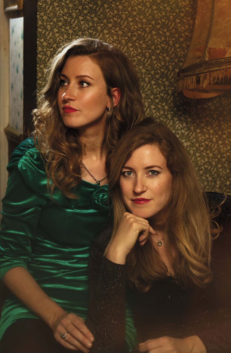 Twee vrouwen poseren samen en glimlachen naar de camera in een warme binnenruimte.