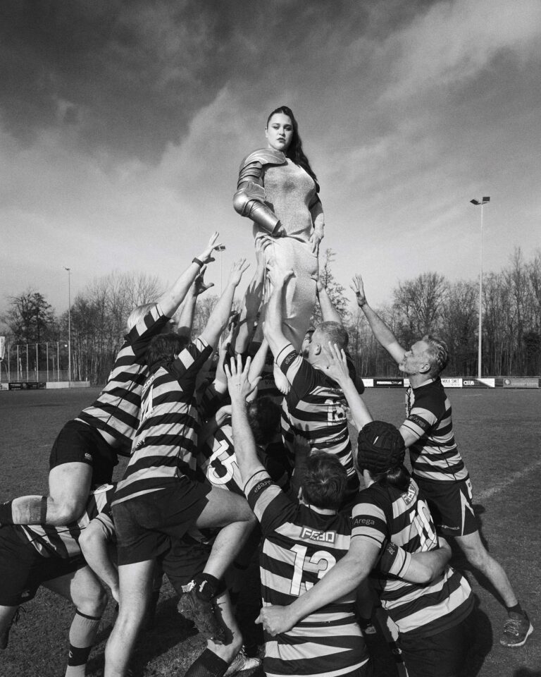 Een vrouw staat op de schouders van meerdere rugbyspelers op een sportveld.