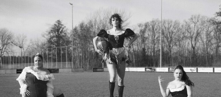 Een persoon poseert in een krachtige houding op een grasveld, gekleed in een donkere top, korte rok en hoge laarzen (zwart-wit foto).